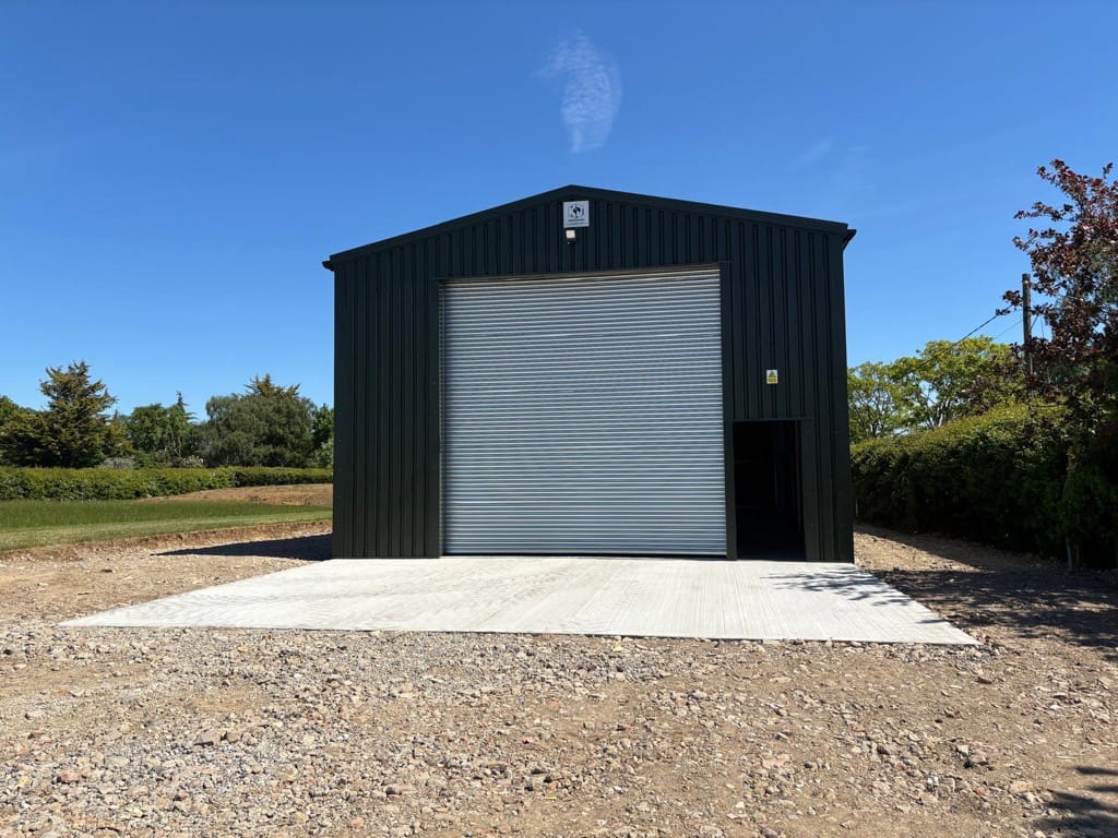 An agricultural steel frame building with a single roller shutter door and a single personnel door to the front gable end. Finished with black cladding.